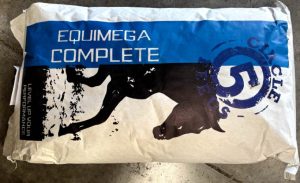 A white bag of Equimega Complete horse feed by Circle 5 Feeds lies on a concrete surface. The bag features a blue and white design with the text "EQUIMEGA COMPLETE" in bold letters and an image of a galloping horse. The Circle 5 Feeds logo, with a large "5" in a circular design, is prominently displayed on the right side. The text "LEVEL UP YOUR PERFORMANCE" is visible on the left.